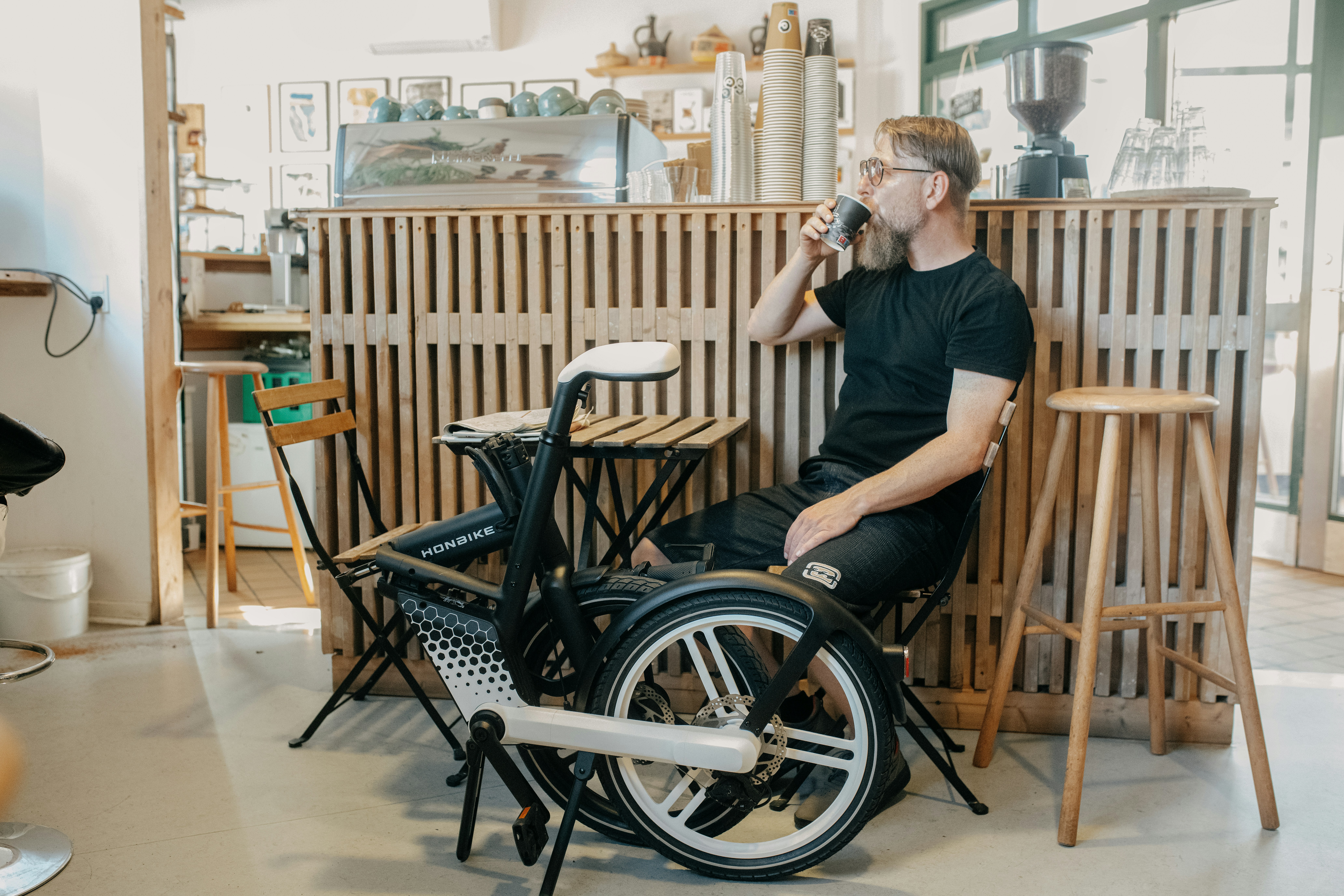 A bearded man sips coffee in a bright, modern cafe with a prominent black and white folding e-bike beside him.