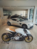 A modern showroom featuring an elegant white motorcycle with golden wheels prominently displayed in the foreground. Behind the motorcycle, several luxury cars, including a sleek silver coupe, are showcased under bright lighting. The showroom has a minimalist design with glass walls and neutral-toned flooring.