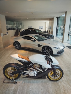A modern showroom featuring an elegant white motorcycle with golden wheels prominently displayed in the foreground. Behind the motorcycle, several luxury cars, including a sleek silver coupe, are showcased under bright lighting. The showroom has a minimalist design with glass walls and neutral-toned flooring.