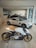 A modern showroom featuring an elegant white motorcycle with golden wheels prominently displayed in the foreground. Behind the motorcycle, several luxury cars, including a sleek silver coupe, are showcased under bright lighting. The showroom has a minimalist design with glass walls and neutral-toned flooring.