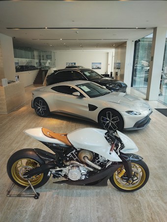 A modern showroom featuring an elegant white motorcycle with golden wheels prominently displayed in the foreground. Behind the motorcycle, several luxury cars, including a sleek silver coupe, are showcased under bright lighting. The showroom has a minimalist design with glass walls and neutral-toned flooring.