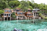 A collection of wooden houses on stilts situated along a pristine, crystal-clear body of water. The houses have large windows and are surrounded by lush green trees. The setting is tranquil and idyllic, with natural rock formations leading into the water.