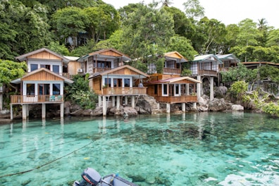 A collection of wooden houses on stilts situated along a pristine, crystal-clear body of water. The houses have large windows and are surrounded by lush green trees. The setting is tranquil and idyllic, with natural rock formations leading into the water.