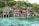 A collection of wooden houses on stilts situated along a pristine, crystal-clear body of water. The houses have large windows and are surrounded by lush green trees. The setting is tranquil and idyllic, with natural rock formations leading into the water.