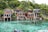 A collection of wooden houses on stilts situated along a pristine, crystal-clear body of water. The houses have large windows and are surrounded by lush green trees. The setting is tranquil and idyllic, with natural rock formations leading into the water.