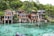 A collection of wooden houses on stilts situated along a pristine, crystal-clear body of water. The houses have large windows and are surrounded by lush green trees. The setting is tranquil and idyllic, with natural rock formations leading into the water.