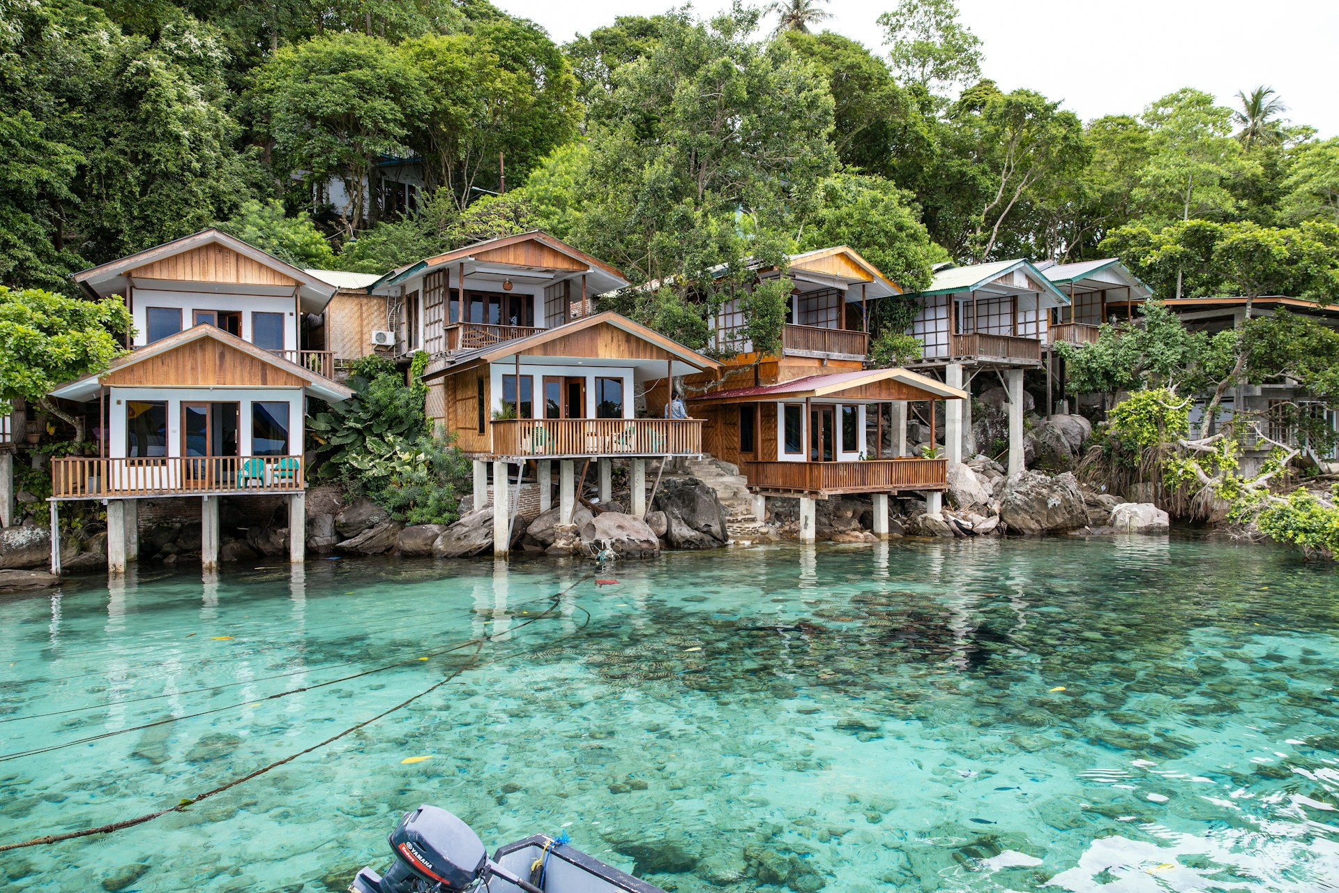 A collection of wooden houses on stilts situated along a pristine, crystal-clear body of water. The houses have large windows and are surrounded by lush green trees. The setting is tranquil and idyllic, with natural rock formations leading into the water.