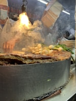 A vendor prepares food in a busy market environment. Steam rises dramatically from a large metal cooking surface filled with various ingredients, such as grilled meat and vegetables. A bright overhead light illuminates the scene, creating an intense focus on the cooking area. The vendor, wearing a mask and apron, is partially visible in the background, adding to the bustling and lively atmosphere.