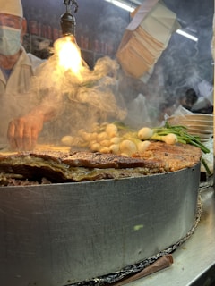 A vendor prepares food in a busy market environment. Steam rises dramatically from a large metal cooking surface filled with various ingredients, such as grilled meat and vegetables. A bright overhead light illuminates the scene, creating an intense focus on the cooking area. The vendor, wearing a mask and apron, is partially visible in the background, adding to the bustling and lively atmosphere.