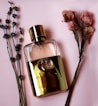 A gold-colored perfume bottle with a prominent logo is placed on a soft pink background, flanked by sprigs of lavender and clusters of dried pink flowers.