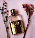 A gold-colored perfume bottle with a prominent logo is placed on a soft pink background, flanked by sprigs of lavender and clusters of dried pink flowers.