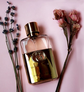 A gold-colored perfume bottle with a prominent logo is placed on a soft pink background, flanked by sprigs of lavender and clusters of dried pink flowers.