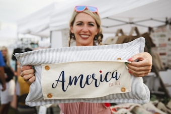 A person is smiling while holding a decorative pillow with the text 'America Est. 1776' printed on it. The pillow has a striped design with buttons, and the background features a market or fair setting with tents and stalls.