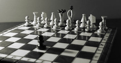 A black chess pawn faces a row of white chess pieces arranged on a board. The background is a neutral gray, subtly lit to enhance the three-dimensionality of the chess pieces. The scene suggests an impending game, with a slightly dramatic composition.