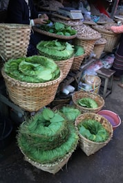 A market scene with neatly stacked green leaves arranged in several woven baskets. The baskets are placed on a wooden stall alongside other containers holding various items like nuts or seeds. The focus is on the vibrant green leaves, with a person seen arranging them, and another person partially visible in the background.