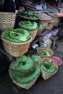 A market scene with neatly stacked green leaves arranged in several woven baskets. The baskets are placed on a wooden stall alongside other containers holding various items like nuts or seeds. The focus is on the vibrant green leaves, with a person seen arranging them, and another person partially visible in the background.