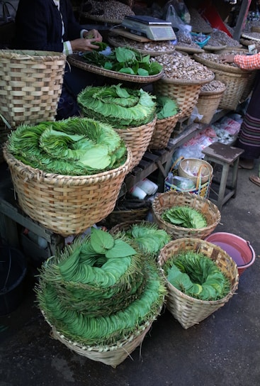 A market scene with neatly stacked green leaves arranged in several woven baskets. The baskets are placed on a wooden stall alongside other containers holding various items like nuts or seeds. The focus is on the vibrant green leaves, with a person seen arranging them, and another person partially visible in the background.