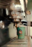 A paper cup with a logo sits on the tray of a coffee machine. The focus is on the cup which has a design featuring an emblem of a horse. The background and parts of the machine are blurred, providing a sense of depth.