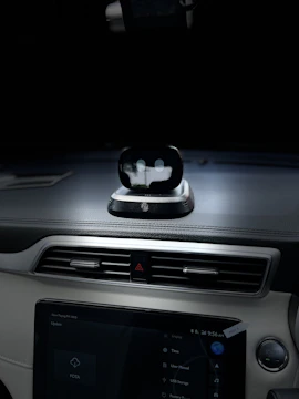 A view of the interior dashboard of a car with a small robot-like device positioned on top of the dashboard. The dashboard also features a touchscreen displaying various options. The environment is dimly lit, focusing on the dashboard area.