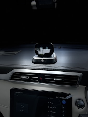 A view of the interior dashboard of a car with a small robot-like device positioned on top of the dashboard. The dashboard also features a touchscreen displaying various options. The environment is dimly lit, focusing on the dashboard area.