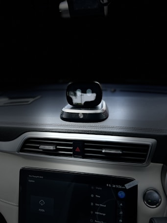 A view of the interior dashboard of a car with a small robot-like device positioned on top of the dashboard. The dashboard also features a touchscreen displaying various options. The environment is dimly lit, focusing on the dashboard area.