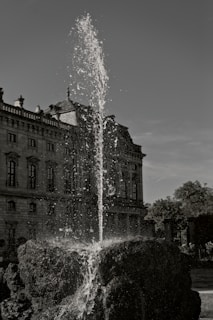 A grand architectural building with ornate details stands in the background, while a tall fountain rises prominently in the foreground. Water cascades down from the fountain, surrounded by rocky formations. The scene includes well-maintained gardens and trees.