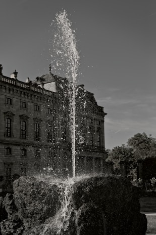 A grand architectural building with ornate details stands in the background, while a tall fountain rises prominently in the foreground. Water cascades down from the fountain, surrounded by rocky formations. The scene includes well-maintained gardens and trees.