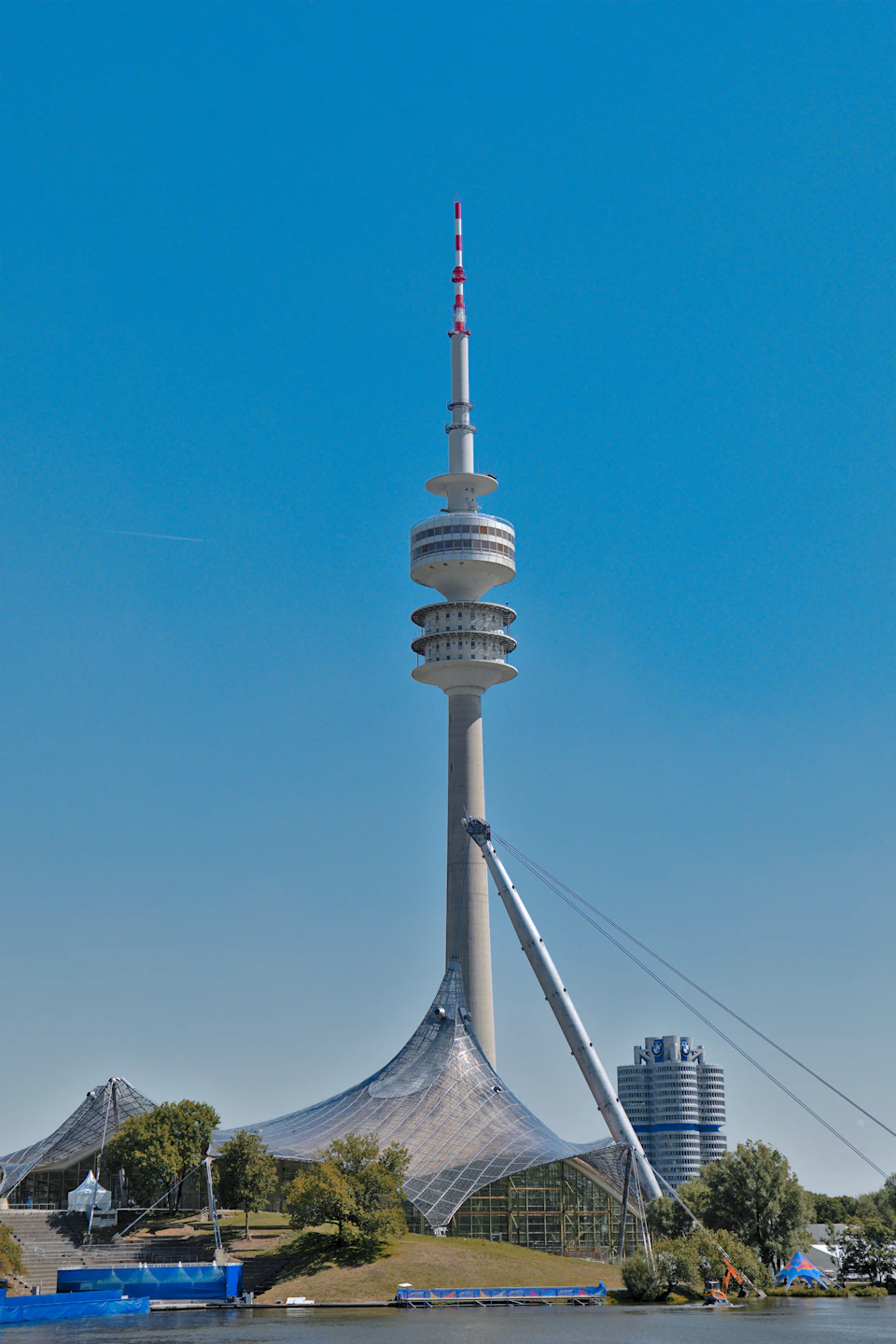 The image shows a large, white tower with multiple levels and antennas on top, standing tall against a clear blue sky. The tower is situated near a body of water, adding to its grandeur. In the foreground, there are two smaller structures - one featuring a curved roof and the other having a flat roof. These structures appear to be part of an architectural project or development in progress.