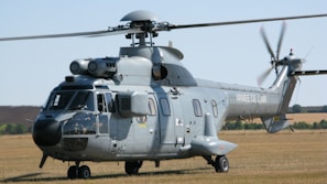 A large, grey military helicopter is parked on a grassy airfield. The helicopter has twin rotors and various equipment mounted on its body. The words 'Armee de l'Air' are visible on the side, indicating it belongs to the French Air Force. The background shows a clear sky and open fields.