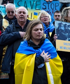 A group of people are gathered, many wrapped in blue and yellow flags, symbolizing Ukrainian support. They appear solemn or reflective, with some holding signs with messages advocating peace and stopping war. The colors blue and yellow are prominent in the scene.