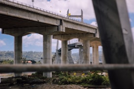 A large concrete bridge spans across the image, supported by several massive pillars. The structure appears to lead across a body of water, with greenery and possibly a town visible in the background under a partly cloudy sky.
