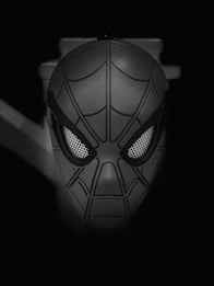 A monochrome image of a superhero mask, featuring prominent web-like patterns and large, expressive eyes. The mask appears to be floating in the darkness, creating a dramatic contrast.