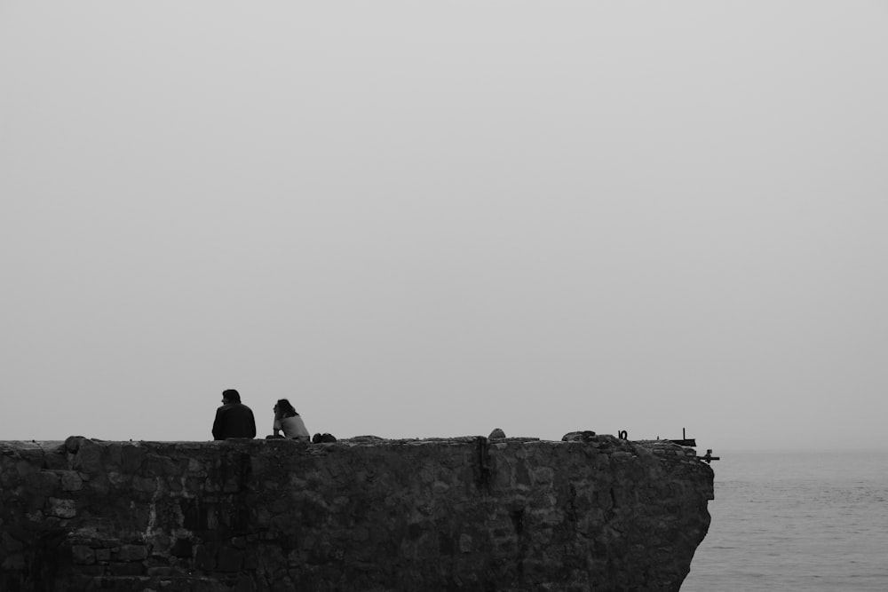 a couple of people sitting on a stone wall overlooking the ocean