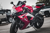 A sleek red and black sports motorcycle is parked on a concrete surface, surrounded by other vehicles in what appears to be a parking lot. The motorcycle is designed with sharp angles and aerodynamic features, including prominent decals and logos.