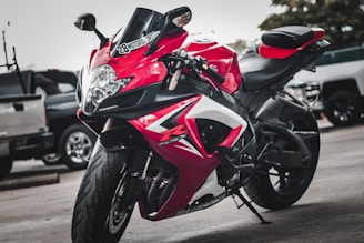 A sleek red and black sports motorcycle is parked on a concrete surface, surrounded by other vehicles in what appears to be a parking lot. The motorcycle is designed with sharp angles and aerodynamic features, including prominent decals and logos.