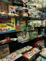 A glass display case filled with vintage toys and board games featuring themes from 1960s and 1970s pop culture, including models, action figures, and games inspired by comics, TV shows, and science fiction. Prominent items include a Doctor Who board game with Daleks, a Batman puzzle, an Avengers themed game, and various model kits.