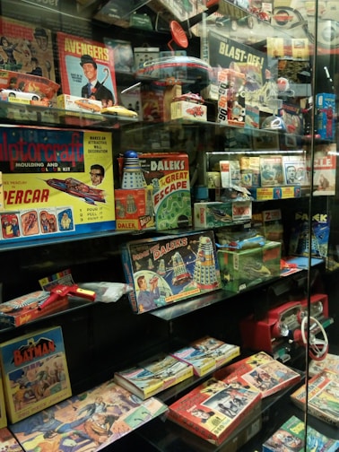 A glass display case filled with vintage toys and board games featuring themes from 1960s and 1970s pop culture, including models, action figures, and games inspired by comics, TV shows, and science fiction. Prominent items include a Doctor Who board game with Daleks, a Batman puzzle, an Avengers themed game, and various model kits.