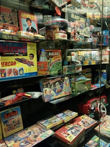 A glass display case filled with vintage toys and board games featuring themes from 1960s and 1970s pop culture, including models, action figures, and games inspired by comics, TV shows, and science fiction. Prominent items include a Doctor Who board game with Daleks, a Batman puzzle, an Avengers themed game, and various model kits.