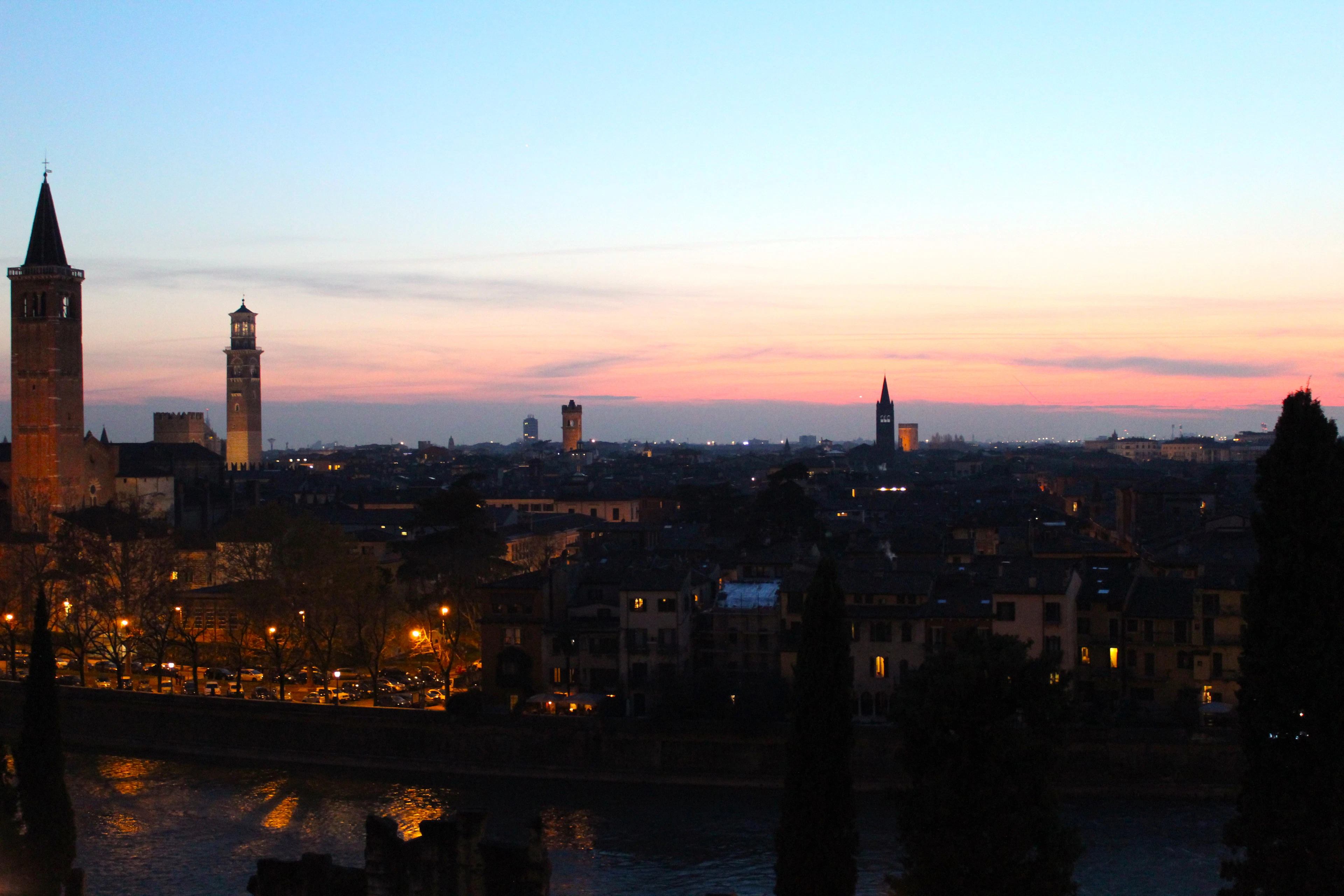 Verona's rooftops and bell towers silhouetted against a pink dusk sky as city lights begin to glow.