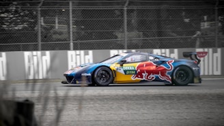 A sleek, high-performance race car with colorful branding and sponsor logos is speeding on a racetrack. The car features prominent Red Bull graphics and is set against a background of a fenced track and blurred surroundings, indicating motion.