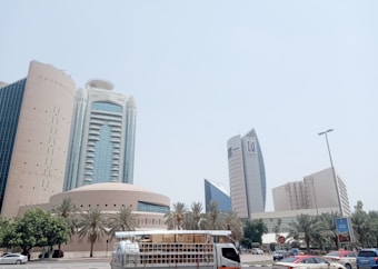 A cityscape with modern skyscrapers featuring unique architectural designs. The scene includes palm trees lining the street and various vehicles on the road, including a white truck in the foreground.