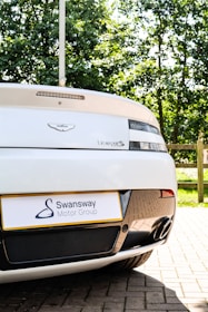 A close-up view of the rear of a white sports car, featuring a visible badge and model name. There is a Swansway Motor Group license plate and two exhaust pipes. The background includes green trees and a wooden fence, suggesting a parking area.