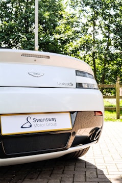 A close-up view of the rear of a white sports car, featuring a visible badge and model name. There is a Swansway Motor Group license plate and two exhaust pipes. The background includes green trees and a wooden fence, suggesting a parking area.
