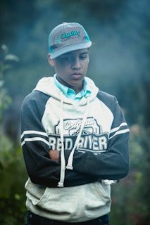 A person wearing a cap with 'Quality Contractors' written on it stands with arms crossed. The person is dressed in a hoodie with the words 'Red River' on the front, layered over a light blue shirt. The background is blurred with greenery, and the focus is on the individual's contemplative expression.