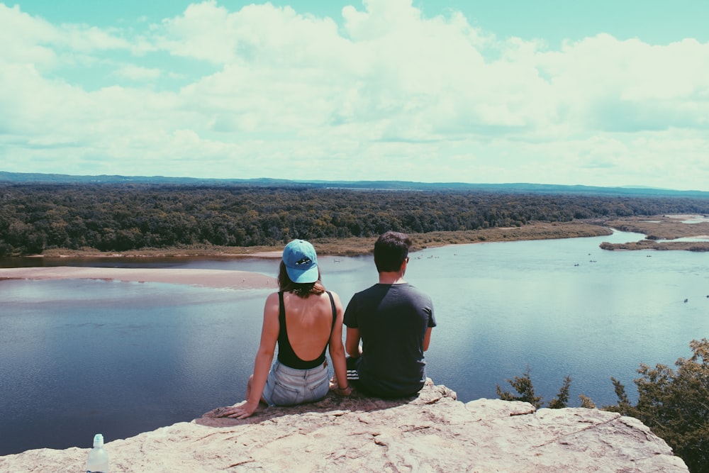 a man and woman sitting on a rock looking at a body of water