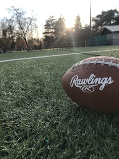 A football rests on a green field with the setting sun creating a warm glow in the background. The field is made of artificial turf and surrounded by trees and some building structures.
