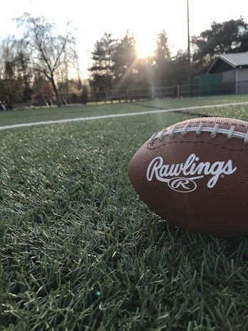 A football rests on a green field with the setting sun creating a warm glow in the background. The field is made of artificial turf and surrounded by trees and some building structures.