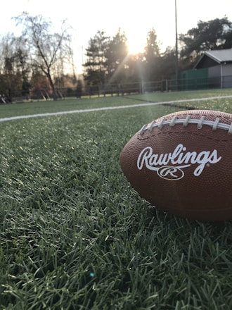 A football rests on a green field with the setting sun creating a warm glow in the background. The field is made of artificial turf and surrounded by trees and some building structures.