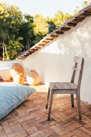 une terrasse entre ombre et soleil avec un grand coussin, une chaise en bois et des pots en terre
