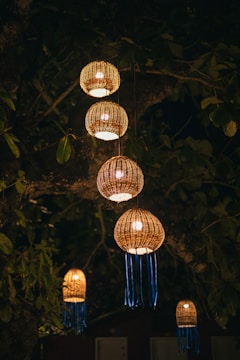 Several woven lanterns hang from tree branches, illuminated against a dark night backdrop. The lanterns emit a warm, yellowish glow, with decorative blue ribbons hanging from their bases. Surrounding leaves and branches create a natural, rustic ambiance.